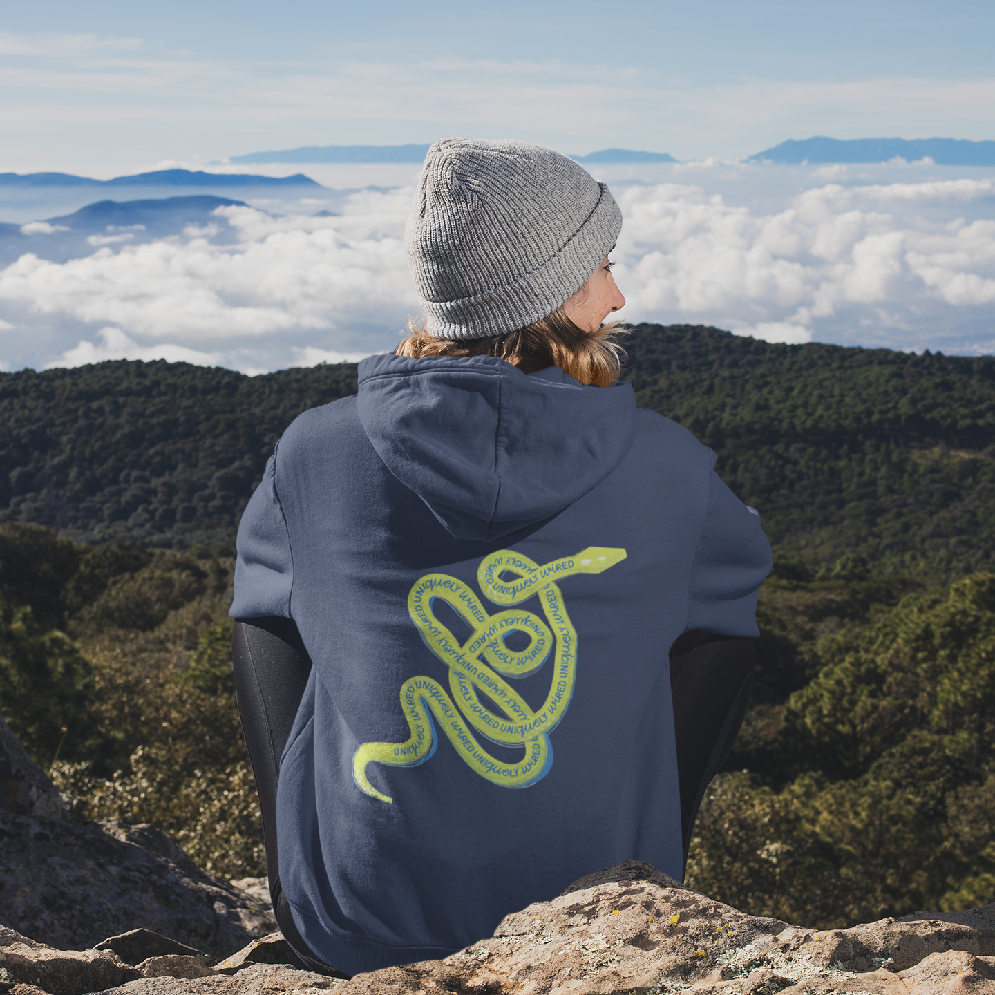 Person wearing a hoodie with a snake design, sitting on a mountain overlooking a cloud-covered valley.