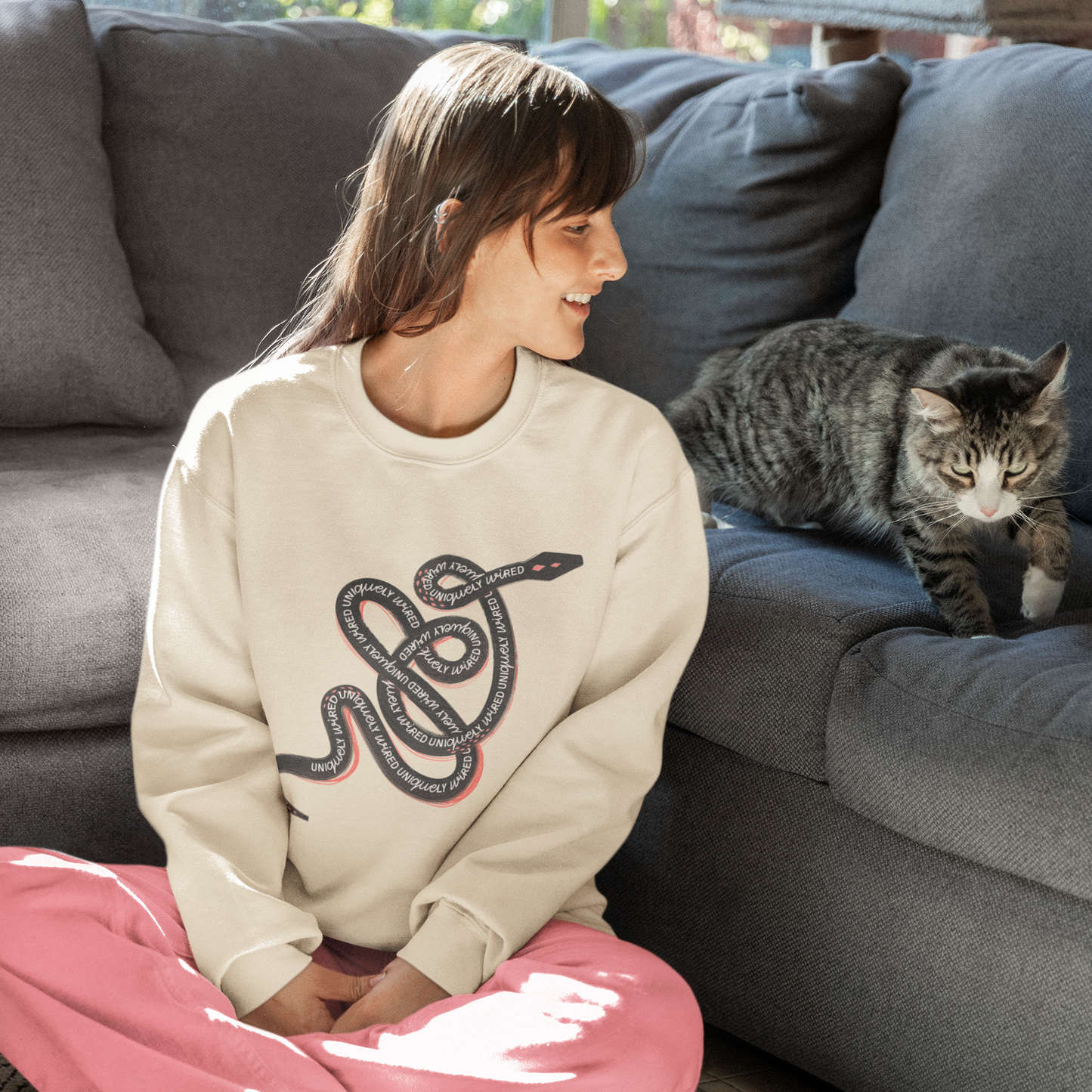 Woman sitting on the floor wearing a beige sweatshirt with a snake design and pink pants, next to a gray cat on a couch.
