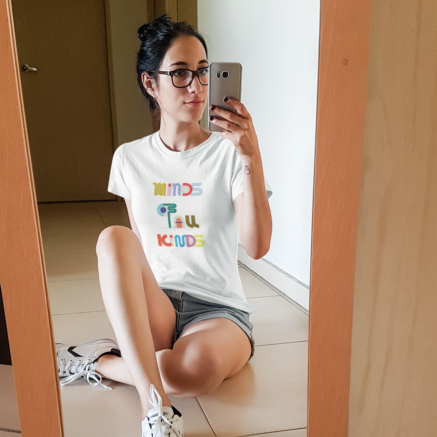 Person taking a mirror selfie wearing a white t-shirt with colorful text and gray shorts.