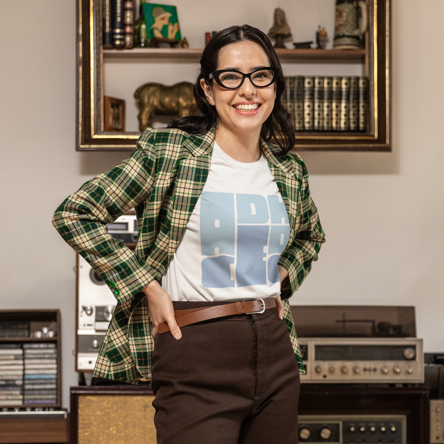 Person wearing a plaid shirt over a graphic t-shirt in a room with vintage record players and books.
