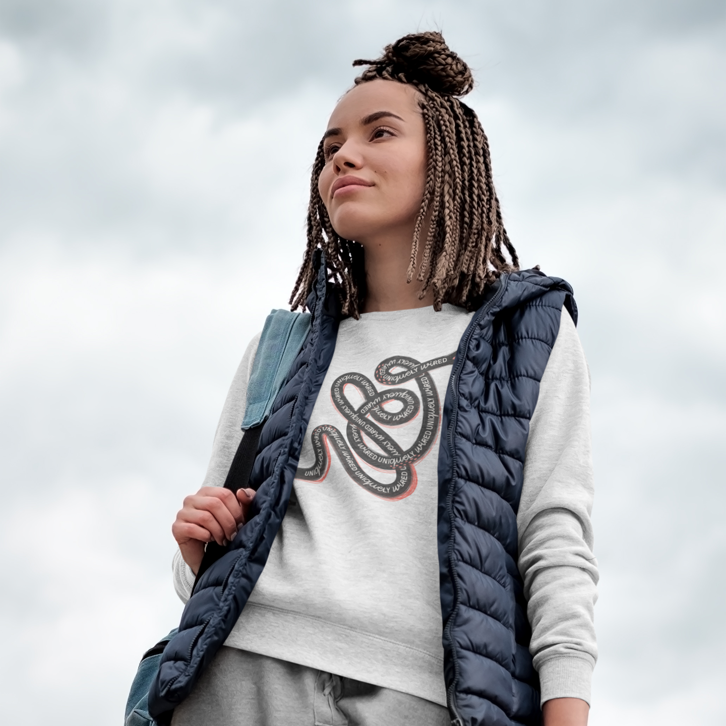 Person wearing a white sweatshirt with a graphic design, gray pants, and a blue puffer vest, holding a skateboard against a cloudy sky.